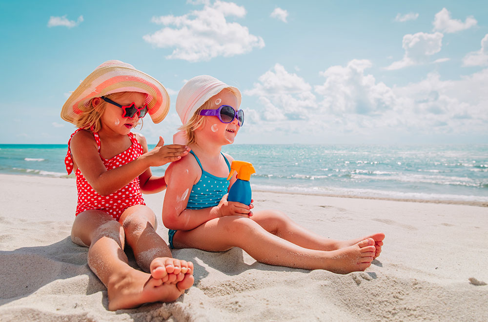 Dvije djevojke na plaži u kupaćim naočalama, sa sunčanim naočalama i šeširima, nanose kremu za sunčanje. - fotografija