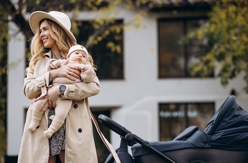 Žena drži bebu u naručju dok hoda pored kolica ispred zgrade.